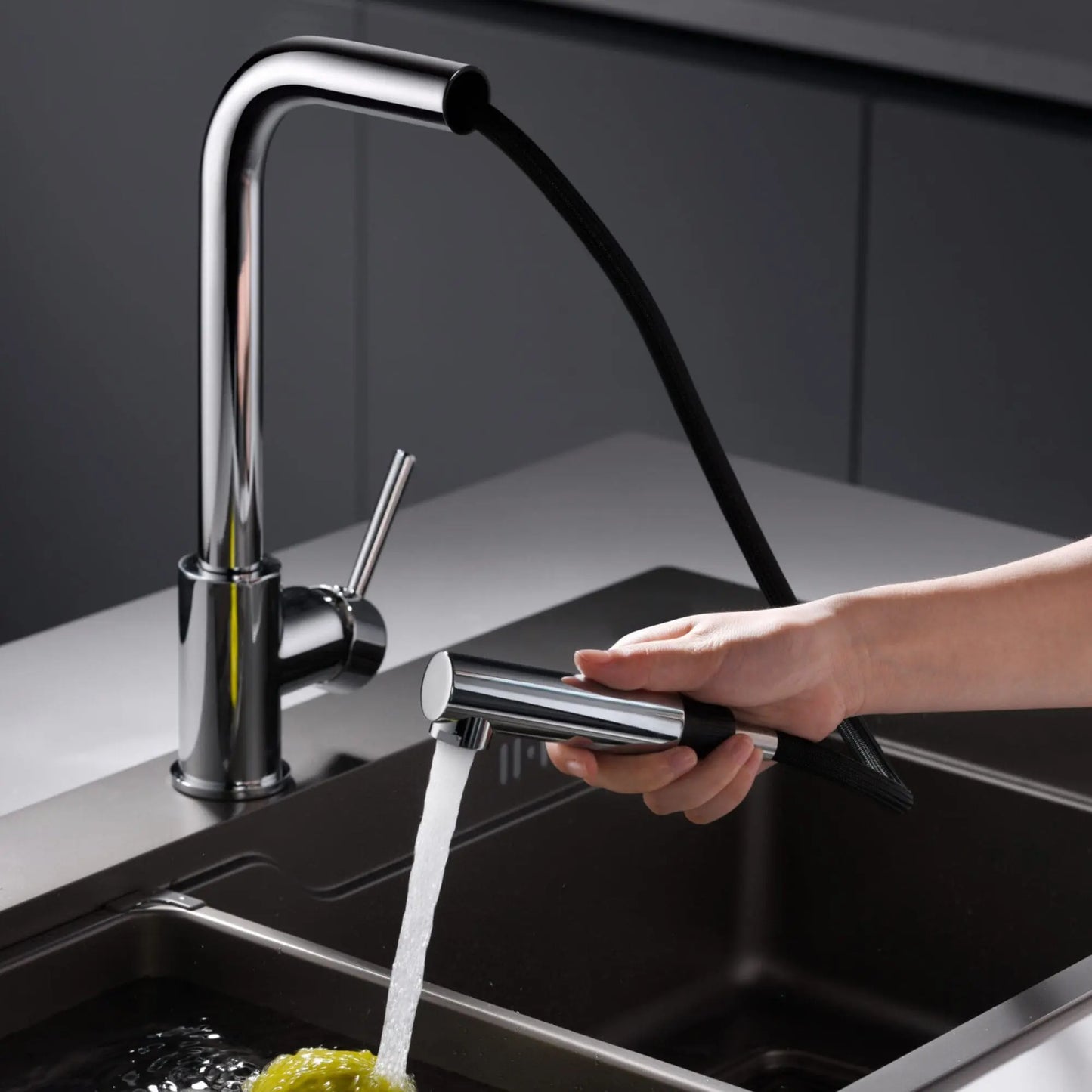 alt='Mão a utilizar o bico extraível da torneira de cozinha Imex Malta para lavar loiça num lava-loiça escuro.'